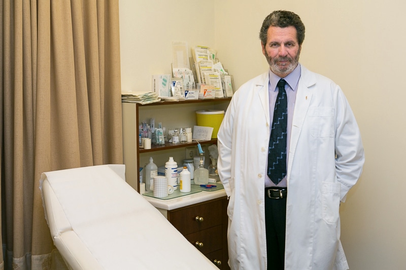 Male doctor in a white lab coat standing beside an exam table in a clinic, with shelves of medical supplies behind him.